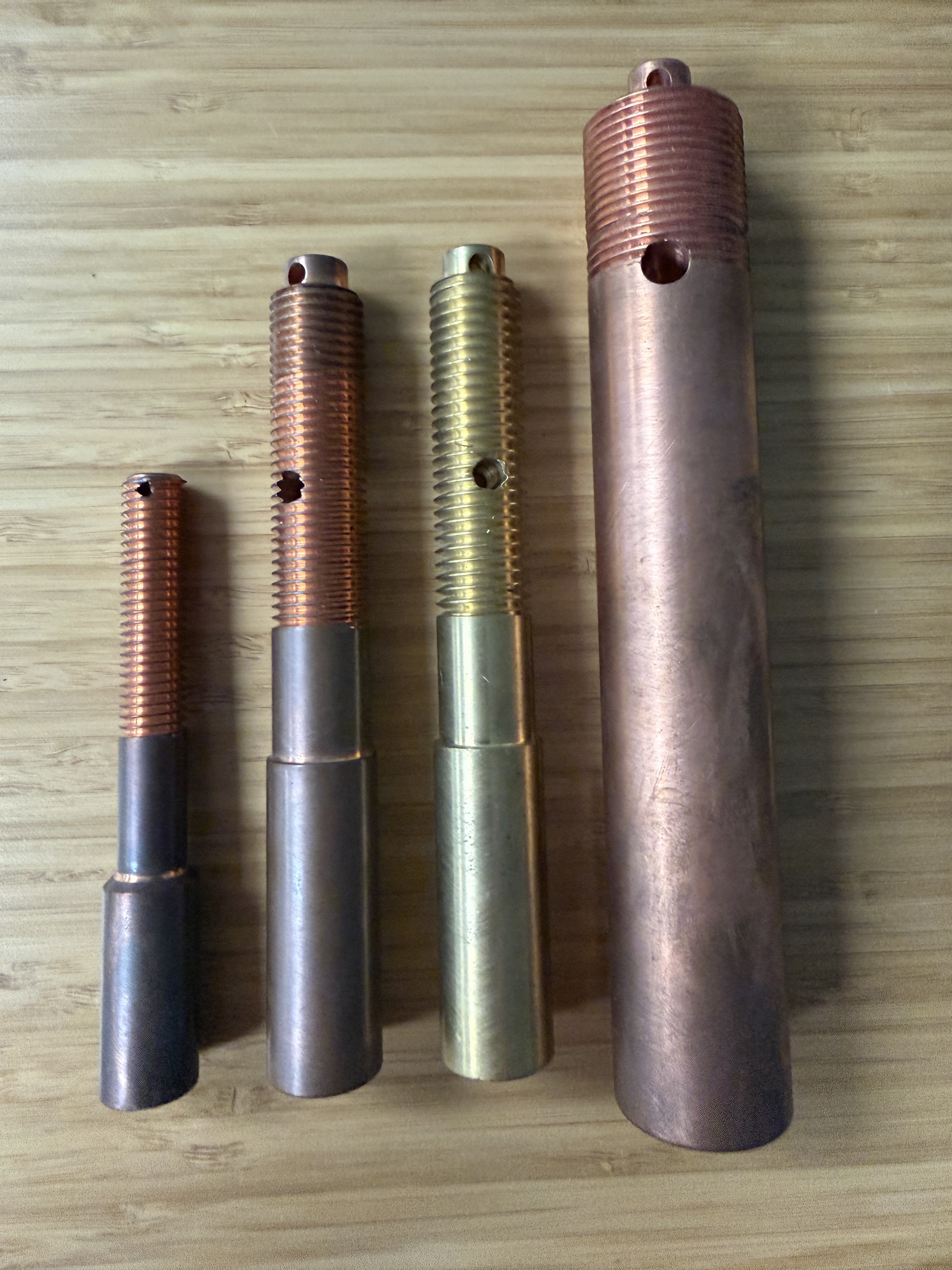 Four transformer bushing draw lead extensions of varying sizes, small steel-bodied copper-threaded, medium copper-threaded with steel shank, medium brass-bodied, and large copper, showing the threaded top ends.